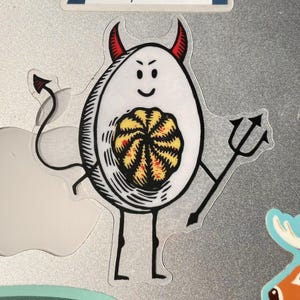 Pode incluir: Um ovo branco com chifres de diabo e uma cauda, segurando um tridente. O ovo tem uma gema amarela e vermelha dentro.