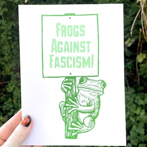 Puede incluir: Una impresión blanca con una rana verde sosteniendo un cartel que dice "FROGS AGAINST FASCISM!". La rana está ilustrada con un estilo de linograbado detallado, y el texto está en negrita, con una fuente sans-serif.