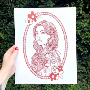 Puede incluir: Una impresión de arte de línea roja de una mujer con cabello largo y fluido, enmarcada por un círculo de cuerda con flores rojas. La impresión está en papel blanco.