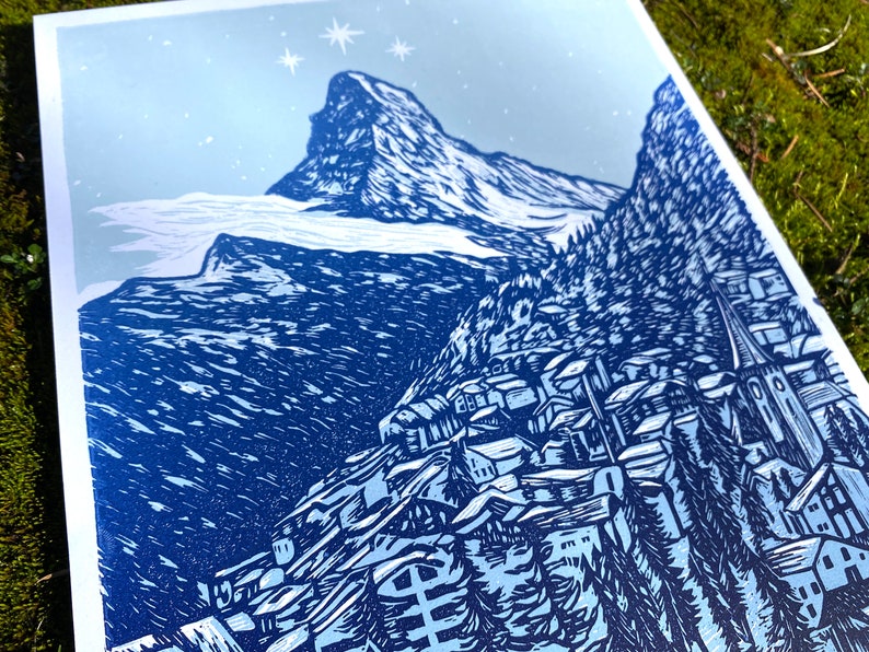 May include: A blue and white woodcut print depicting a mountain village with a snow-capped mountain peak in the background. The print has a vintage, rustic feel.