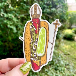 Könnte beinhalten: Ein Aufkleber mit einem Cartoon-Hotdog, der als Papst verkleidet ist, komplett mit Mitra und Stab. Der Hotdog hat ein lächelndes Gesicht und ist mit Relish und einer Gurke gefüllt. Der Aufkleber befindet sich vor einem Hintergrund aus grünem Laub.