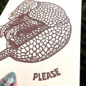 以下が含まれることがあります： 「LEAVE ME ALONE」と「PLEASE」という茶色の文字が書かれた、ベージュ色のカード。アルマジロの詳細な茶色のイラストが描かれています。緑色のマニキュアを塗った人がカードを持っています。
