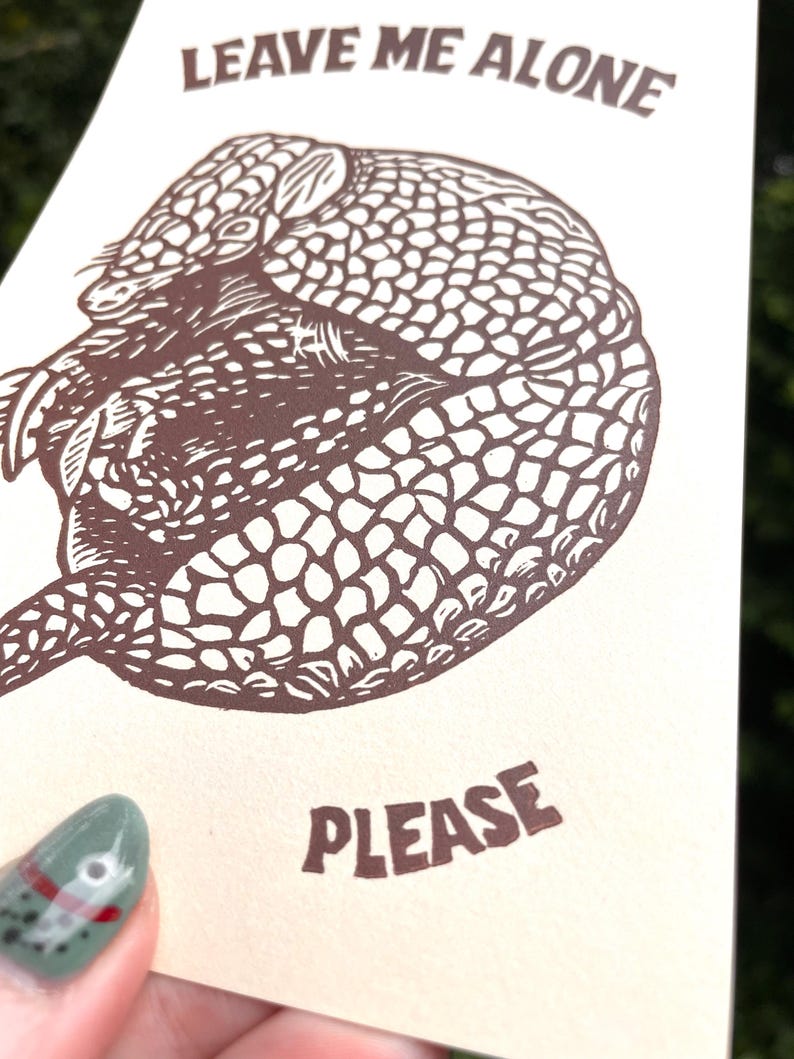 以下が含まれることがあります： 「LEAVE ME ALONE」と「PLEASE」という茶色の文字が書かれた、ベージュ色のカード。アルマジロの詳細な茶色のイラストが描かれています。緑色のマニキュアを塗った人がカードを持っています。