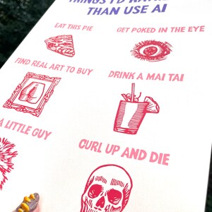 May include: A cream-colored print with red illustrations and text. The text reads "THINGS I'D RATHER DO THAN USE AI." The illustrations include a slice of pie, an eye, a framed artwork, a cocktail, a flower, and a skull.