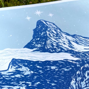 May include: A blue and white linocut print of a mountain peak with a starry sky above. The mountain is depicted in a stylised, blocky style, with sharp peaks and ridges. The sky is a light blue, with white stars scattered across it.