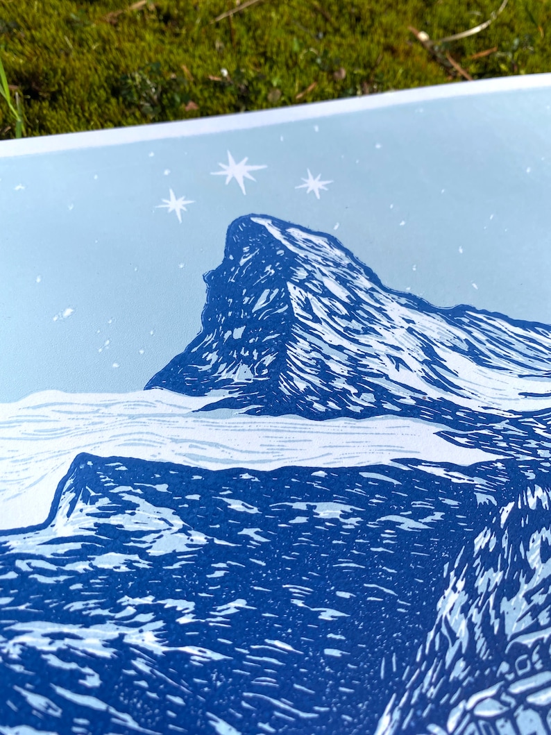 May include: A blue and white linocut print of a mountain peak with a starry sky above. The mountain is depicted in a stylised, blocky style, with sharp peaks and ridges. The sky is a light blue, with white stars scattered across it.