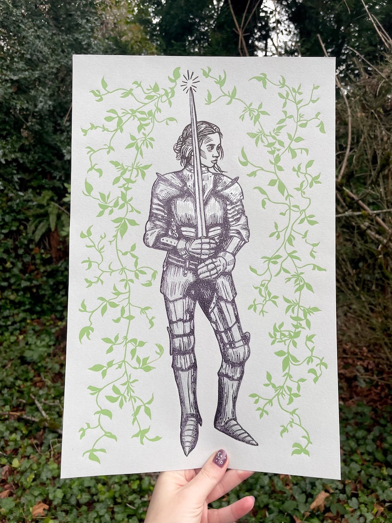 May include: A black and white illustration of a woman in armour holding a sword. The background is a light green with a vine pattern.