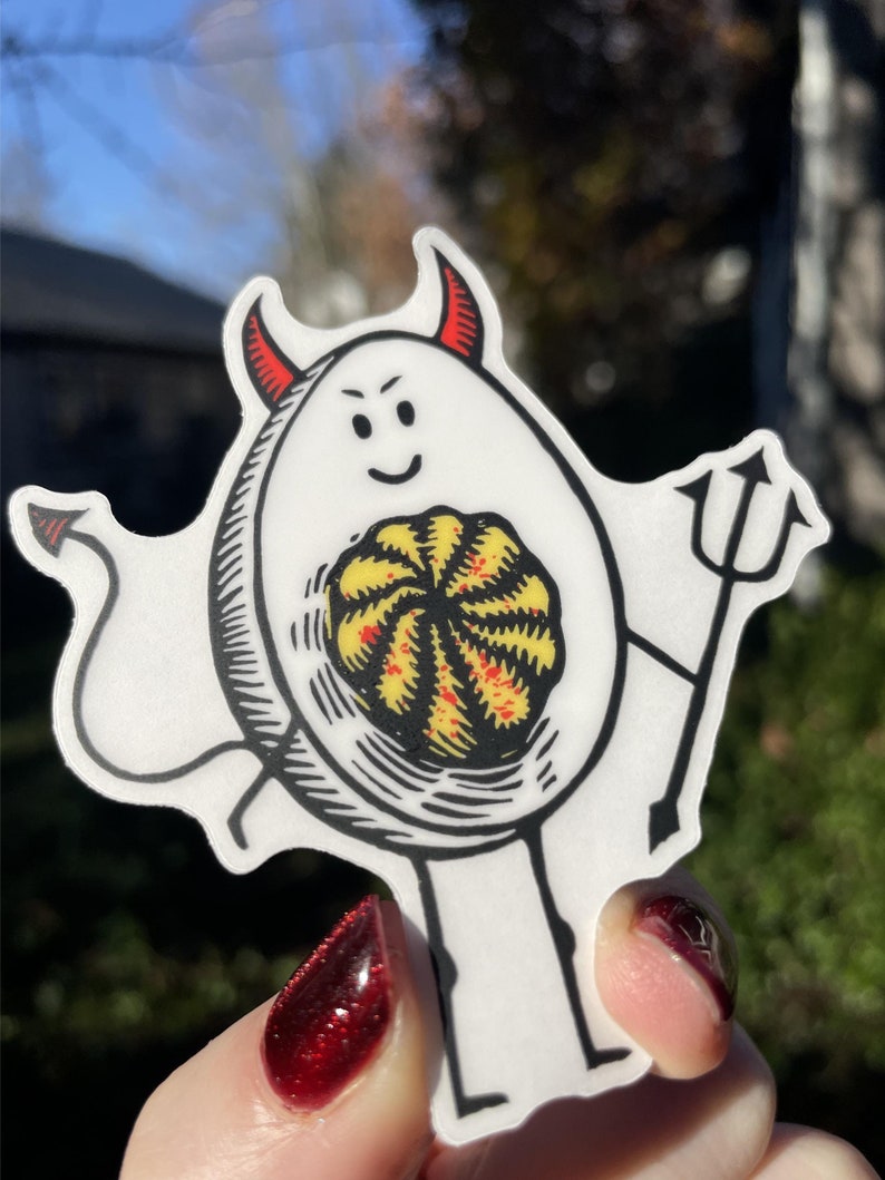 Pode incluir: Um ovo branco com uma fantasia de diabo, chifres vermelhos e uma cauda. O ovo tem um rosto sorridente e est&aacute; segurando um forcado. A gema do ovo &eacute; vis&iacute;vel e tem um padr&atilde;o de redemoinho vermelho e amarelo.