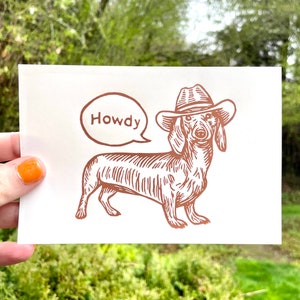 Peut inclure: Un teckel brun et blanc portant un chapeau de cowboy brun avec le mot "Howdy" dans une bulle de dialogue au-dessus de lui.