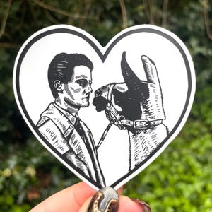Puede incluir: Una pegatina en forma de corazón en blanco y negro que representa a un hombre y una llama mirándose. El hombre lleva un traje y corbata. La llama tiene un cabestro. La pegatina tiene un borde blanco.