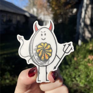Pode incluir: Um adesivo branco com uma ilustra&ccedil;&atilde;o de desenho animado de um diabo com chifres vermelhos e um tridente. O diabo est&aacute; segurando um padr&atilde;o de redemoinho amarelo e vermelho em suas m&atilde;os.