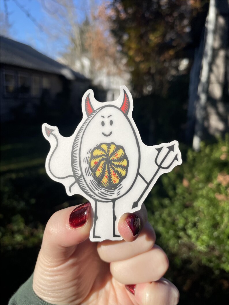 Pode incluir: Um adesivo branco com uma ilustra&ccedil;&atilde;o de desenho animado de um diabo com chifres vermelhos e um tridente. O diabo est&aacute; segurando um padr&atilde;o de redemoinho amarelo e vermelho em suas m&atilde;os.