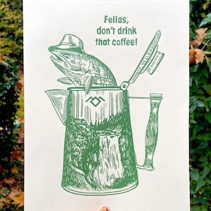 Puede incluir: Impresión verde que representa un pez con sombrero saliendo de una cafetera. El texto dice "Fellas, don't drink that coffee!". La cafetera tiene un diseño de paisaje. La impresión está sobre un fondo crema.