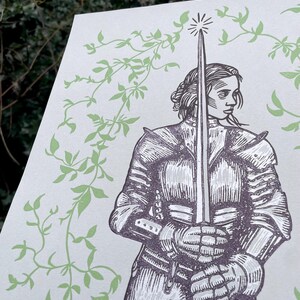 May include: A black and white woodcut print of a woman in armour holding a sword. The woman is standing in front of a background of green vines.