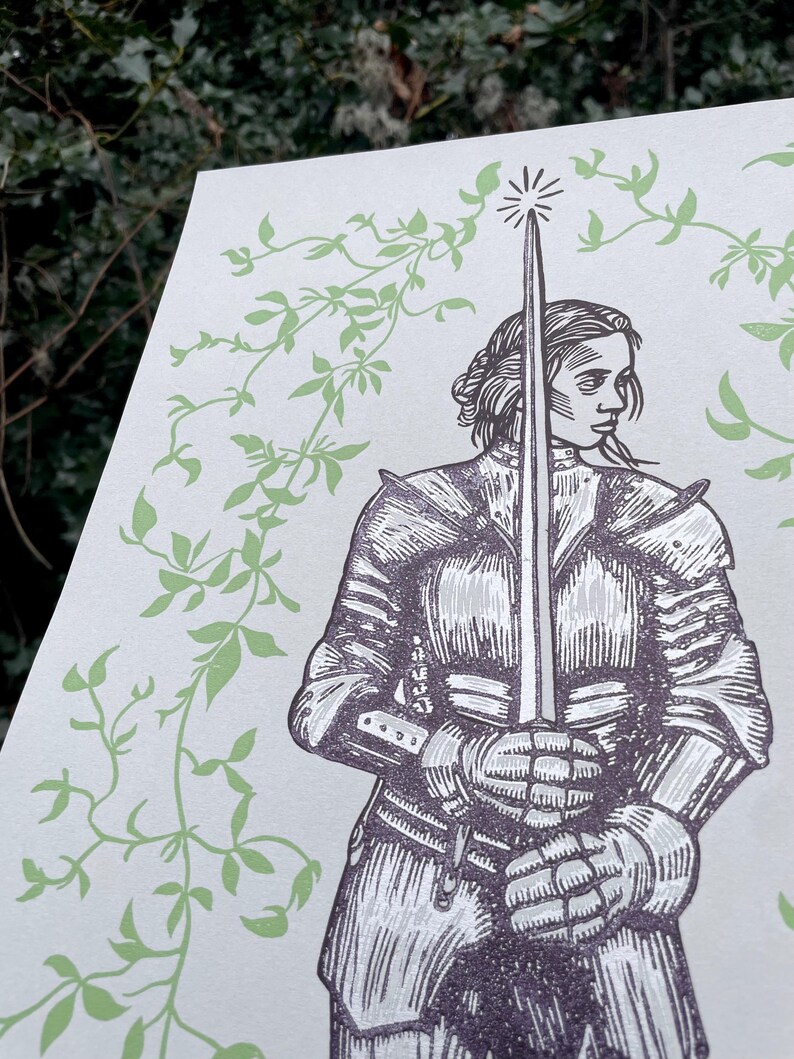 May include: A black and white woodcut print of a woman in armour holding a sword. The woman is standing in front of a background of green vines.