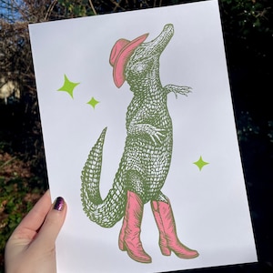 May include: A green alligator wearing a pink cowboy hat and pink cowboy boots. The alligator is standing on a white background with green stars.