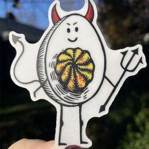 Pode incluir: Um ovo branco com chifres de diabo e um tridente, segurando uma gema amarela e vermelha dentro. O ovo tem um contorno preto e uma cara sorridente.