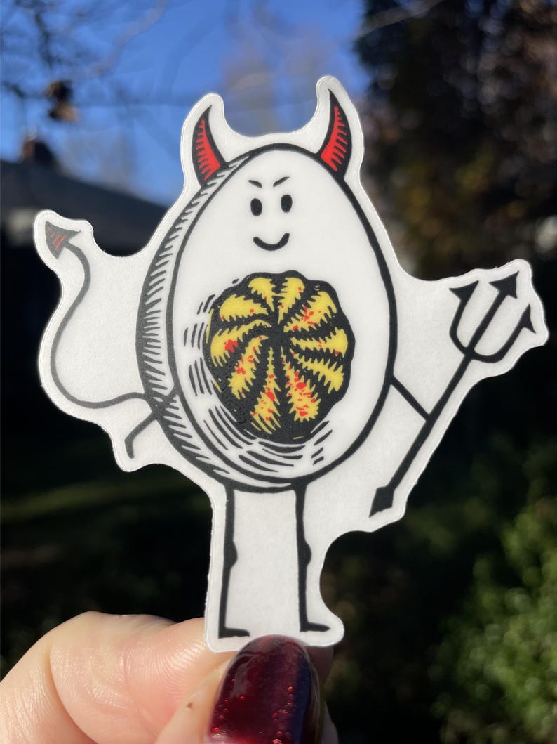 Pode incluir: Um ovo branco com chifres de diabo e um tridente, segurando uma gema amarela e vermelha dentro. O ovo tem um contorno preto e uma cara sorridente.