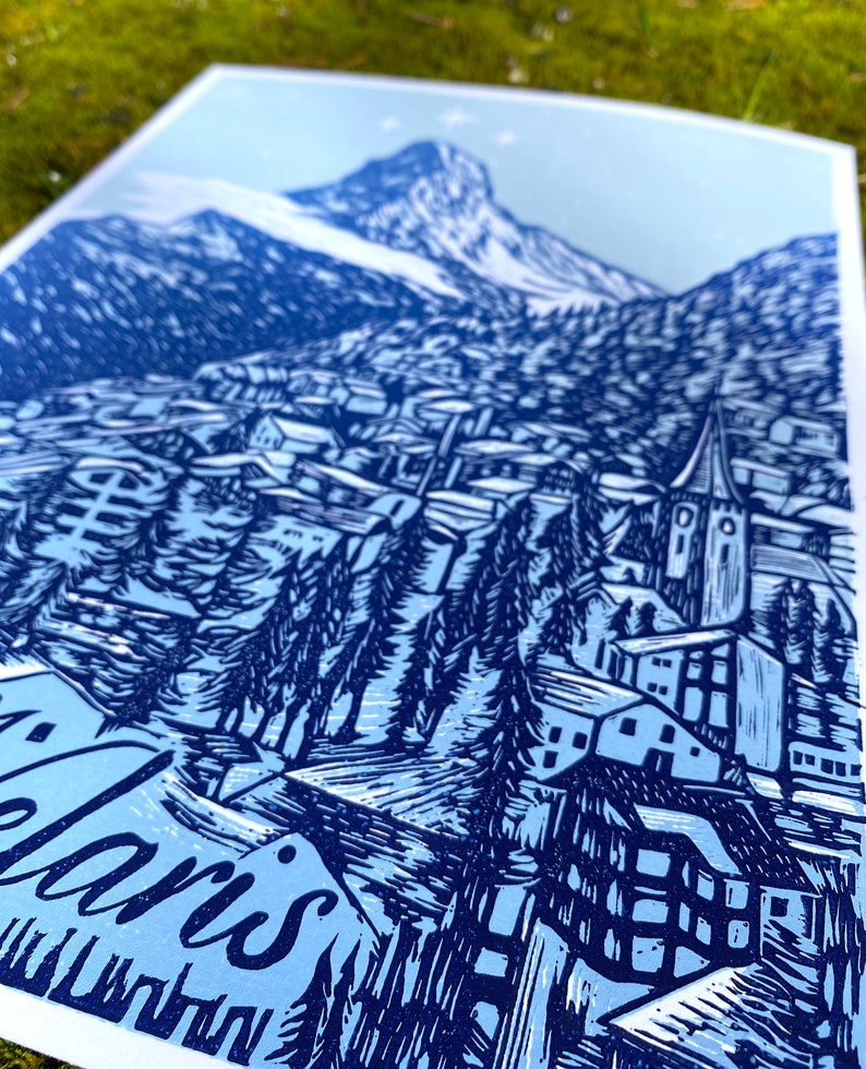 May include: A blue and white linocut print of a mountain village with a church steeple and the text "Alaris" at the bottom.