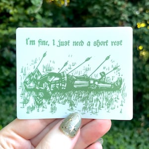 May include: A rectangular white sticker featuring a green ink illustration of a knight lying on the ground with arrows protruding from their armor. The text "I'm fine, I just need a short rest" is printed in a green gothic-style font above the knight.