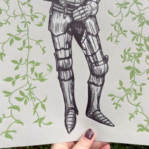 May include: A black and white illustration of a knight in armour standing in front of a green leafy background.