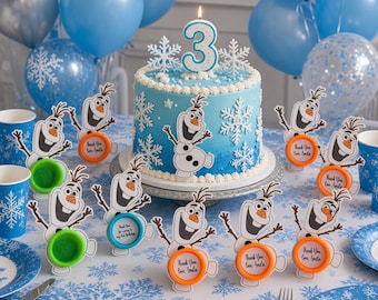 May include: A blue ombre birthday cake topped with a number 3 candle, surrounded by Olaf character party favors with orange and green circular bases, placed on a snowflake-patterned tablecloth with blue and silver balloons in the background.