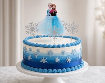 May include: A round cake decorated with blue ombre frosting, silver snowflake cutouts, and a blue tulle skirt topper featuring two small figures. The cake has blue piped borders at the top and bottom edges and sits on a white pedestal stand.