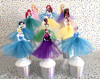 May include: A collection of cupcake toppers featuring various animated princess characters, each wearing a layered, glittery tulle skirt in colors including blue, green, yellow, purple, and white, set atop silver metallic cupcake liners.