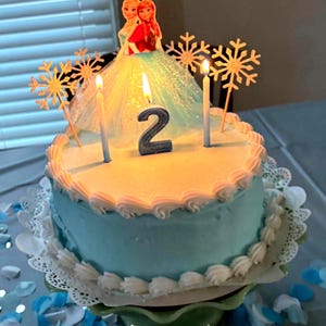 May include: A blue and white frosted cake with a number 2 candle and two Frozen characters on top. The cake is decorated with snowflake toppers and is on a white doily.