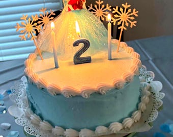Puede incluir: Un pastel glaseado azul y blanco con una vela con el número 2 y dos personajes de Frozen en la parte superior. El pastel está decorado con adornos de copo de nieve y está sobre un encaje blanco.
