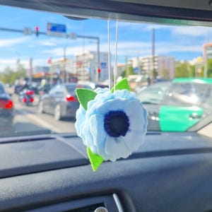 Pode incluir: Um ambientador de carro em forma de flor de feltro azul claro, com um centro azul escuro e folhas de feltro verdes, pendurado no espelho retrovisor. O painel do carro e uma cena de rua movimentada são visíveis no fundo.