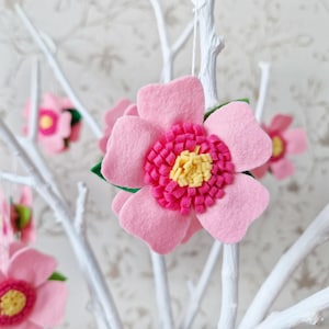 以下が含まれることがあります： 白い枝から吊るされた、明るいピンク色の中心と黄色のアクセントが付いたピンク色のフェルトフラワーオーナメント。花には緑の葉があり、花のディスプレイのように配置されています。