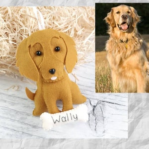 Puede incluir: Un adorno de perro de fieltro con un hueso marr&oacute;n y blanco, el nombre "Wally" est&aacute; bordado en el hueso. El adorno est&aacute; sobre una superficie de madera blanca. Un golden retriever aparece en el fondo.