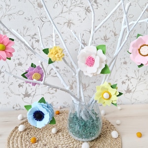 Può includere: Un ramo bianco con ornamenti di fiori in feltro di vari colori, tra cui rosa, giallo, blu e viola. I fiori sono appesi ai rami e sono disposti in un vaso.