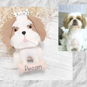 Puede incluir: Un perro de juguete de fieltro blanco con orejas marrones, un lazo blanco y negro y una lengua rosa. El perro sostiene un hueso rosa con el nombre "Pecan" bordado.