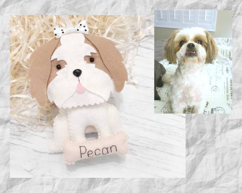 Puede incluir: Un perro de juguete de fieltro blanco con orejas marrones, un lazo blanco y negro y una lengua rosa. El perro sostiene un hueso rosa con el nombre "Pecan" bordado.