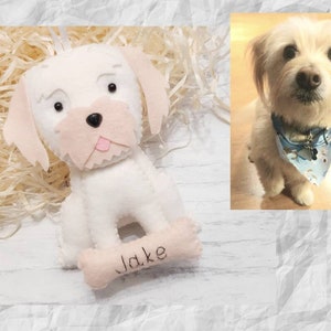 Puede incluir: Adorno de perro de fieltro blanco con ojos negros y nariz rosa. El adorno tiene una etiqueta en forma de hueso con el nombre "Jake" escrito en ella.