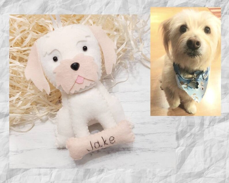 Puede incluir: Adorno de perro de fieltro blanco con ojos negros y nariz rosa. El adorno tiene una etiqueta en forma de hueso con el nombre "Jake" escrito en ella.