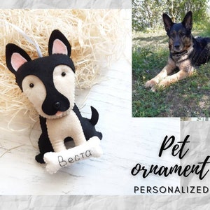 Puede incluir: Un adorno de perro de fieltro personalizado con pelaje blanco y negro, sosteniendo un hueso con el nombre "Besta" bordado. El adorno est&aacute; colgado de una cuerda blanca. La imagen tambi&eacute;n muestra un perro real, un pastor alem&aacute;n, tumbado en la hierba.