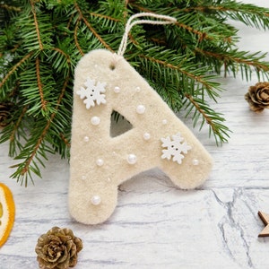 Può includere: Un ornamento natalizio in feltro beige a forma di lettera "A", decorato con fiocchi di neve bianchi e piccole perle. L'ornamento è appeso a un filo ed è ambientato su uno sfondo di rami di pino, stelle di legno e una fetta d'arancia essiccata.