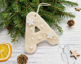 May include: A beige felt Christmas ornament in the shape of the letter "A", decorated with white snowflakes and small pearls. The ornament hangs from a string and is set against a backdrop of pine branches, wooden stars, and a dried orange slice.