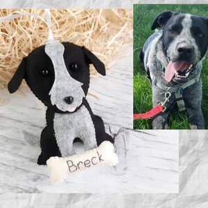 Puede incluir: Un adorno de fieltro de un perro blanco y negro con una mancha gris en la cara. El adorno sostiene un hueso con el nombre "Breck" bordado.