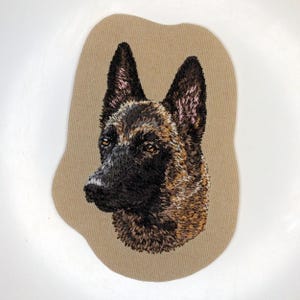 May include: Embroidered patch depicting a dog's head, with detailed stitching in brown, black, and tan. The dog's fur and facial features are carefully rendered. The patch is set against a tan background, creating a striking contrast.