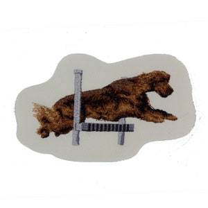 May include: Embroidered patch of a brown dog jumping over a gray hurdle. The dog is in mid-air, with its legs extended and its head tilted forward. The hurdle is a simple, two-bar design.