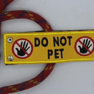 Può includere: Toppa in tessuto giallo con bordo nero e cuciture gialle. La toppa ha due cerchi rossi con una mano nera con una linea diagonale che la attraversa. Il testo "DO NOT PET" è in nero.