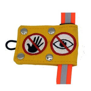 Peut inclure: Pochette d'entraînement pour chien en toile jaune avec une boucle élastique noire, deux boutons-pression en métal argenté et deux cercles rouges brodés avec une main noire barrée d'une barre oblique et un œil noir barré d'une barre oblique. La pochette est attachée à une sangle orange vif avec une bande réfléchissante argentée.