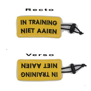 May include: Yellow fabric dog training tag with black text that reads "IN TRAINING NIET AAIEN" on one side and "IN TRAINING NIET AAIEN" on the other side. The tag has a black elastic loop.
