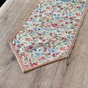 Pode incluir: Um corredor de mesa floral com uma borda festonada e um fundo bege. O corredor tem um padr&atilde;o floral em tons de vermelho, azul e verde. Uma pequena planta em vaso est&aacute; em cima do corredor de mesa.