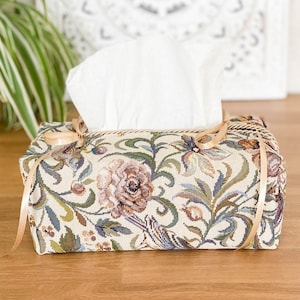 Può includere: Un copri scatola di fazzoletti beige con un design a tappezzeria floreale con uccelli e fiori. Il copri scatola ha un nastro dorato legato intorno.