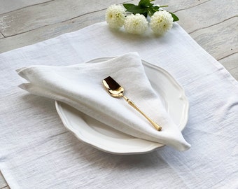 May include: A white linen placemat with a white linen napkin folded on top of a white ceramic plate. A gold spoon is placed on top of the napkin. White flowers are in the background.